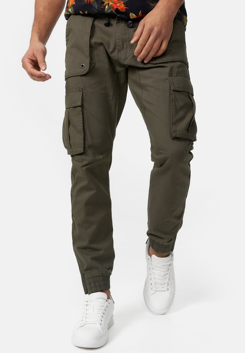 Pantalons cargo vert olive avec des poignets élastiques, plusieurs poches latérales et un tissu léger, associés à des baskets blanches.