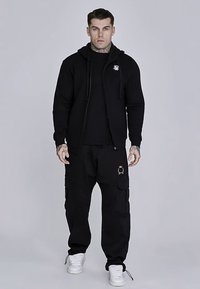 Zwarte hoodie met ritssluiting en logo, gecombineerd met zwarte cargobroek met zakken en gouden accenten. Witte sneakers maken de outfit compleet.