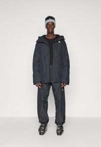 Schwarze wasserdichte Jacke mit Kapuze, kombiniert mit passenden wasserdichten Hosen und Skischuhen. Merkmale sind Logo-Akzente und hochwertiges, langlebiges Material.