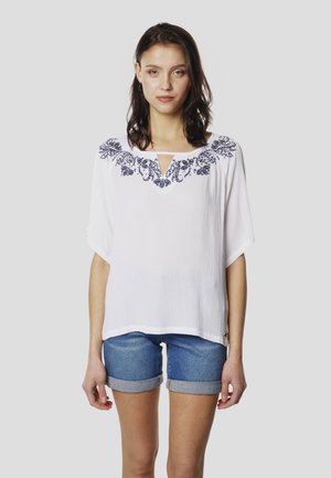 Top blanco de manga corta con acabado texturizado, que presenta un bordado floral azul alrededor del escote y un detalle de abertura en forma de gota en la parte frontal.