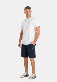 Camiseta blanca de algodón con un pequeño logo en el pecho, combinada con pantalones cortos de carga azul marino con bolsillos laterales. Calzado casual completa el look.