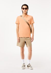 Camisa polo naranja con cuello, pantalones cortos beiges, calcetines blancos y zapatillas bicolor. El modelo sostiene un bolso cruzado marrón y lleva gafas de sol.