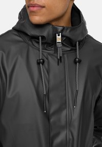 Chaqueta negra impermeable con un alto cuello, que presenta cordones grises y un cierre delantero con cremallera. Textura suave con detalles de diseño minimalistas.