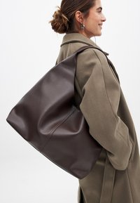 Brun läderhandväska med en avslappnad design, stor öppning och en enda rem, bärs över en olivgrön kappa. Slät textur och minimalistisk stil.