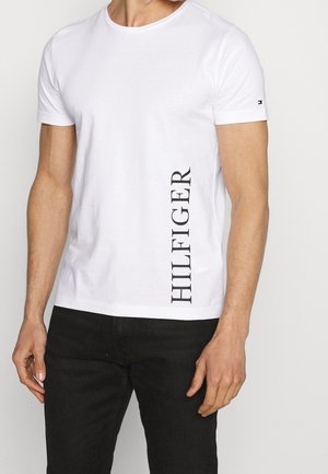 Homme portant un t-shirt blanc à manches courtes avec "HILFIGER" écrit verticalement sur le côté, associé à un pantalon noir.