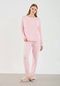 Νεαρή γυναίκα που στέκεται φορώντας ροζ μακρυμάνικο μπλουζάκι και ταιριαστά παντελόνια, με μπεζ αφράτα παντόφλες σε απλό ανοιχτό φόντο.