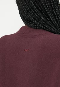 Bordo Nike sweatshirt ima visok ovratnik in mehak material, z majhnim vezenim logotipom na hrbtni strani v usklajeni bordo barvi.