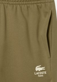 Pantalones cortos de sudadera color verde oliva hechos de tela suave, con cintura elástica y un bolsillo lateral. Logo blanco de Lacoste impreso en la pierna.