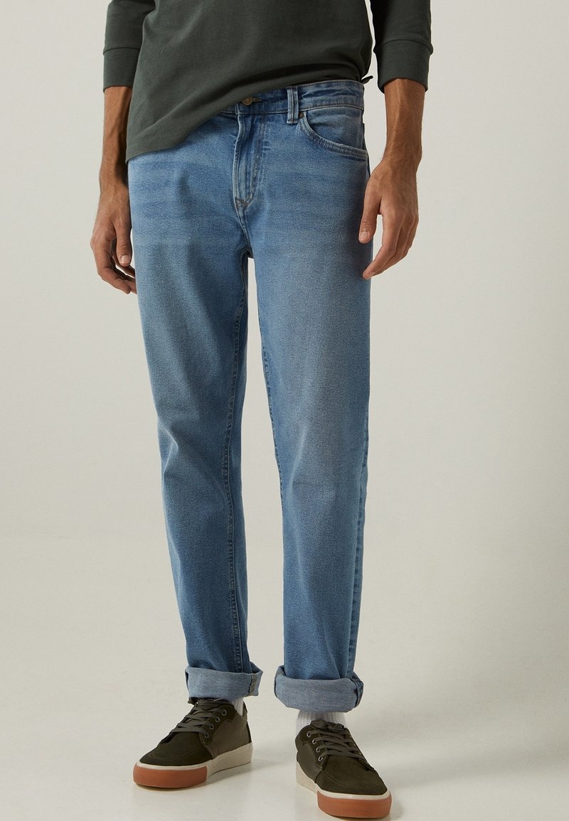 Homme portant un jean bleu clair retroussé, des chaussettes blanches, des baskets vert olive et un haut à manches longues sombre, debout devant un fond uni.