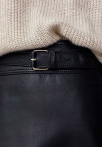 Pull en maille beige glissé dans une ceinture en cuir noir avec boucle dorée, sur un vêtement en cuir noir assorti.