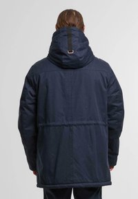 Navy blue puffer jacket with a large hood, featuring a contrasting black strap at the back, and a smooth, textured finish.