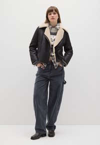 Giacca nera in pelle con colletto in ecopelliccia color crema, indossata sopra un maglione a fantasia e una camicia a quadri, abbinata a pantaloni larghi in velluto a coste scuro.