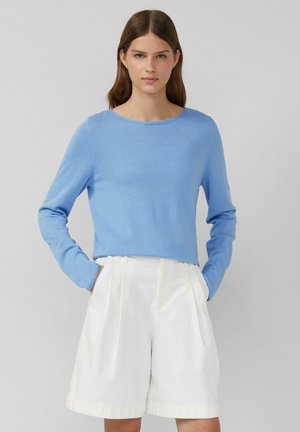 Maglietta azzurra chiara a maniche lunghe con scollatura a girocollo, abbinata a pantaloni corti bianchi con pieghe. Il tessuto sembra morbido e leggermente testurizzato.