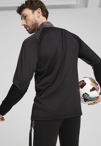 Svart langermet treningsskjorte med grå krage, glatt tekstur og løs passform; holdt fotball med fargerike mønstre og logoer.