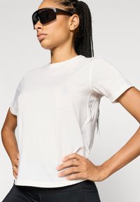 Αθλητική μπλούζα σε ανοιχτό μπεζ χρώμα με κοντά μανίκια, στρογγυλή λαιμόκοψη και διακριτικές ραφές, σε συνδυασμό με κομψά, γωνιώδη μαύρα γυαλιά ηλίου.