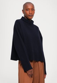 Łysy mężczyzna ubrany w luźny, ciemnoniebieski sweter z golfem oraz wysokie, szerokie brązowe spodnie na jednolitym tle.