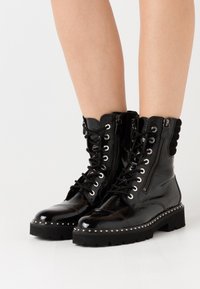 Bottes de combat en cuir verni noir avec laçage sur le devant, fermeture éclair latérale, détails cloutés le long de la semelle et accents texturés en haut.