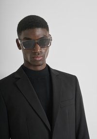 Mlađi muškarac nosi sunčane naočale s prozirnim okvirom, crni sako i crni rolak na jednobojnoj svijetlosivoj pozadini.