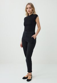 Une femme se tient avec confiance dans un haut noir ajusté et un pantalon noir, les mains dans les poches, sur un fond clair.