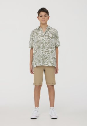 Maglietta verde a maniche corte con motivo di foglie bianche, pantaloni corti beige arrotolati e sneakers bianche. Design semplice, materiali leggeri e vestibilità rilassata.