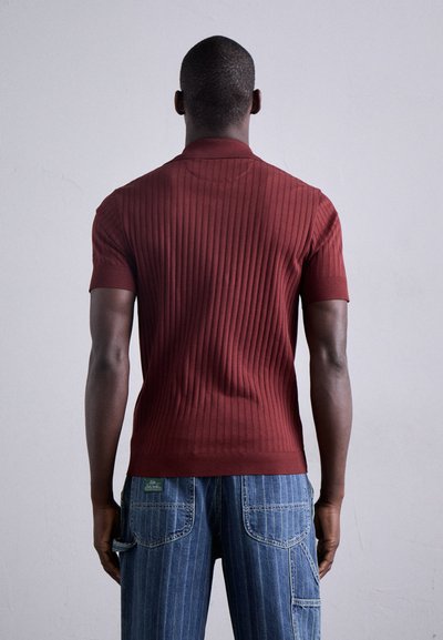Polo shirt bourgogne à manches courtes et côtelé avec une coupe ajustée. Porté avec un jean en denim bleu présentant des rayures verticales et des poches arrière.
