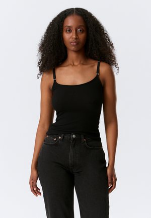 Vrouw met krullend haar, gekleed in een zwart mouwloos topje en zwarte jeans met hoge taille, kijkend naar voren tegen een eenvoudige lichte achtergrond.