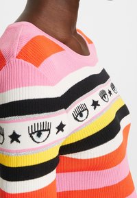 Personne portant un pull rayé rose, orange, jaune, noir et blanc avec des motifs d'œil et d'étoile sur la poitrine et le haut du bras.