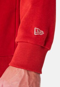 Felpa rossa in cotone con maniche caratterizzate da polsini a coste e un logo ricamato in bianco. La texture è liscia con un pattern di maglia coerente.
