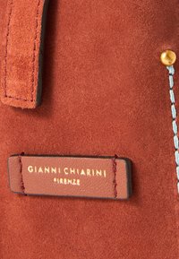 Brun semsket stoff med teksturert overflate. En lærmerking med gullbokstaver er festet, som viser "Gianni Chiarini Firenze."
