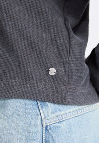 Haut gris texturé avec un tissu côtelé, orné d'un petit logo argenté sur l'ourlet, associé à un denim bleu clair.