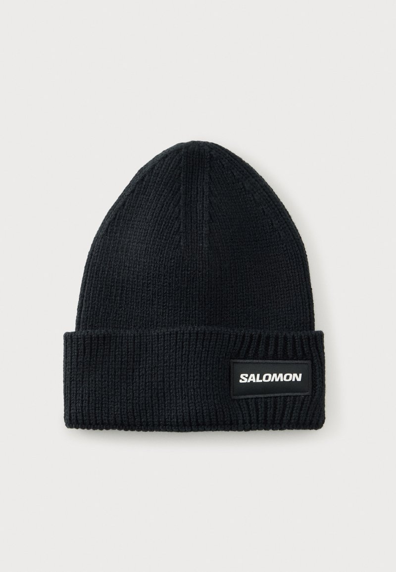 Bonnet noir en tricot côtelé avec revers, comportant un écusson rectangulaire marqué "Salomon" sur fond clair.
