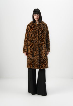 Vrouw staat tegen een witte muur, gekleed in een lange bruin-zwart luipaardprint bontjas en zwarte wijde broek.
