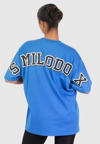Camiseta azul de corte oversize hecha de algodón. Presenta un gran texto negro "SMILODOX" en la espalda y un acento negro en la manga.