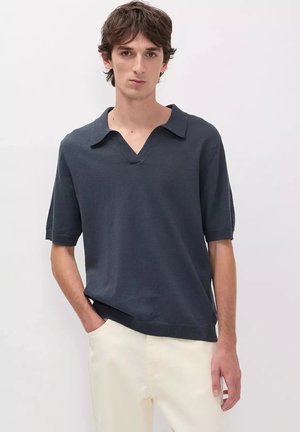 Joven con cabello oscuro de longitud media que lleva una camiseta de punto de manga corta gris oscuro y pantalones beige claros sobre un fondo liso.