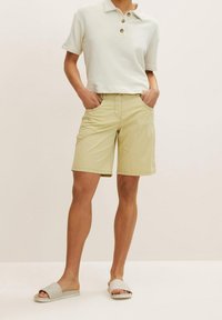 Shorts kaki avec une coupe décontractée, deux poches latérales et une taille à boutons, associés à un polo court blanc et des sandales de couleur claire.