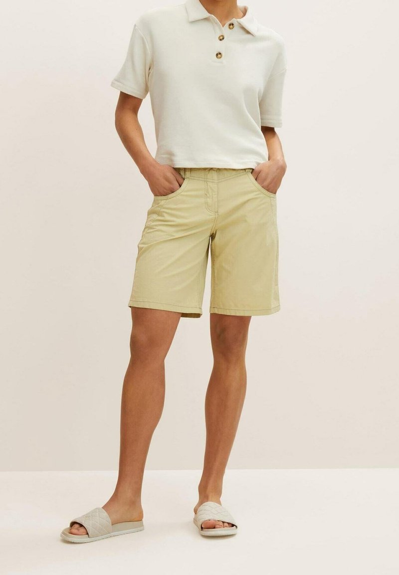Shorts kaki avec une coupe décontractée, deux poches latérales et une taille à boutons, associés à un polo court blanc et des sandales de couleur claire.