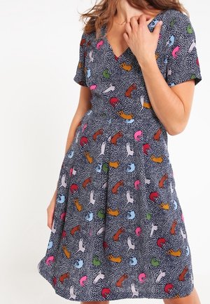 Kobieta ubrana w sukienkę do kolan z krótkim rękawem, ozdobioną kolorowymi, abstrakcyjnymi wzorami dłoni na ciemnym, kropkowanym tle.