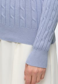 Manche et bas de pull en maille torsadée bleu clair avec poignets côtelés sur une personne portant une jupe ou un pantalon blanc.