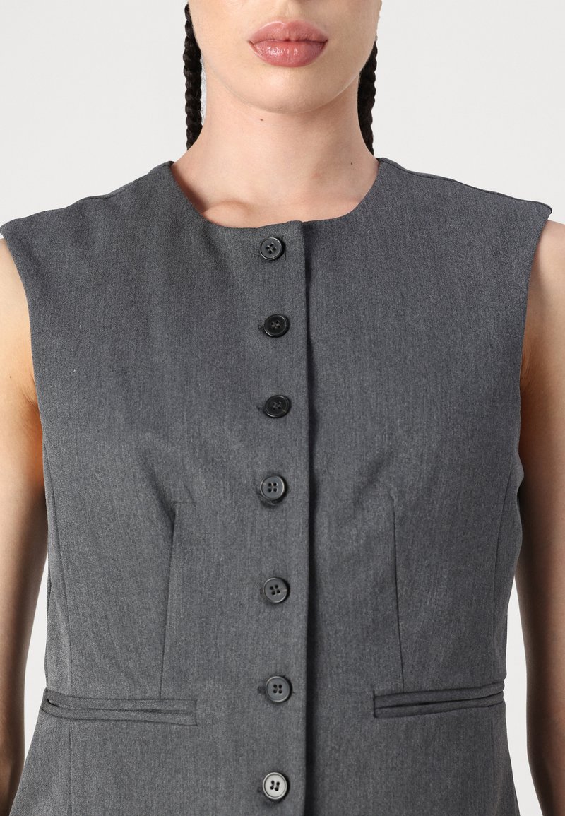 Graues ärmelloses Kleid mit einem runden Ausschnitt, das vorne mit sechs schwarzen Knöpfen und zwei Seitentaschen versehen ist. Weicher, strukturierter Stoff.