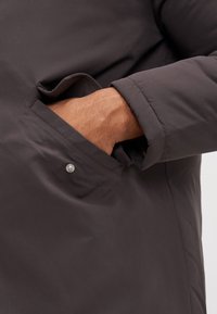 Mörkgrå kappa med en sidficka med en silverknapp och en manschettärm, som visar en slät, matt yta.