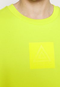 Gros plan d'une personne portant une chemise jaune vif avec un patch logo carré affichant un triangle et le mot "ICEBERG" sur la poitrine.