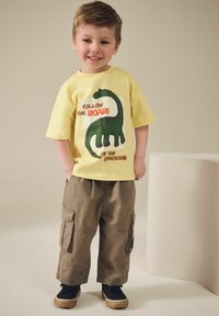 Gul kortærmet t-shirt med grøn dinosaur grafik og tekst, parret med løstsiddende khaki cargo bukser og sorte sneakers.