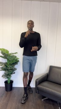 Schwarzes Langarmshirt, Denim-Shorts, schwarze Schuhe mit Socken. Steht vor einer Topfpflanze und einem schwarzen Ledersofa an einer weißen Wand.