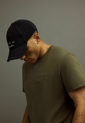 Oliwkowy t-shirt z wytłoczonym tekstem, w zestawieniu z czarną czapką z logo i akcentami, na neutralnym tle.