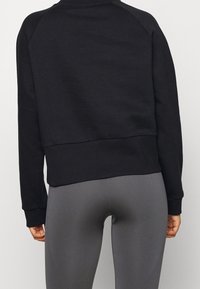 Persona vestida con una camiseta de manga larga negra y leggings ajustados grises, vista de la cintura hacia abajo y de espaldas contra un fondo liso.