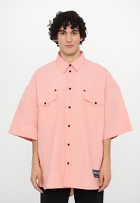 Camisa de manga corta de gran tamaño en tela de color rosa claro, con botones negros y dos bolsillos en el pecho. Incluye una etiqueta de la marca en el lateral.