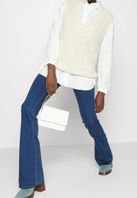 Beige stickad ärmlös tröja, vit skjorta under, blå utsvängda jeans, mintgröna klackstövlar, vit handväska med kedja.