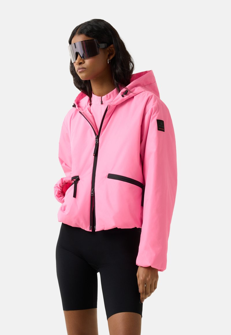 Frau trägt eine übergroße rosa Kapuzenjacke mit schwarzen Reißverschlussdetails, schwarze Radfahrshorts und große reflektierende Visier-Sonnenbrillen.