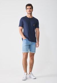 Navy blue cotton t-shirt with a small yellow logo on the pocket, paired with light blue denim shorts, and white sneakers.