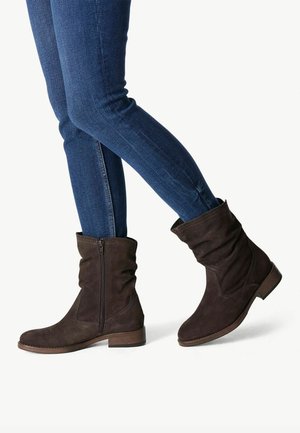 Dunkelbraune Wildlederstiefel bis zur Mitte der Wade mit seitlichem Reißverschluss, getragen mit eng anliegenden blauen Jeans, auf hellem Hintergrund dargestellt.
